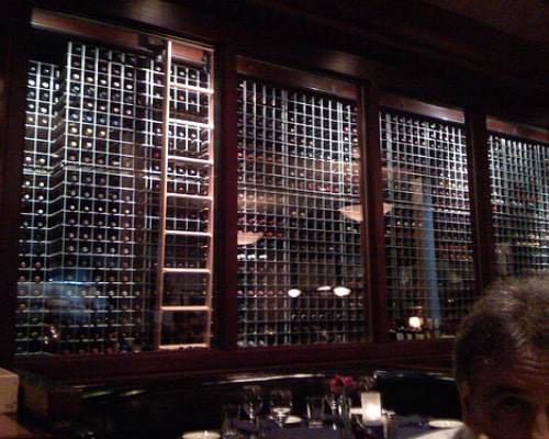 Wall of wine bottles in a restaurant, viewed through glass panels and displayed on elegant wine racks for bars and clubs, with a dining table in the foreground